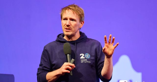 Nicolas Grekas with a mic in his right hand raising his left hand on stage at SymfonyCon Amsterdam 2025