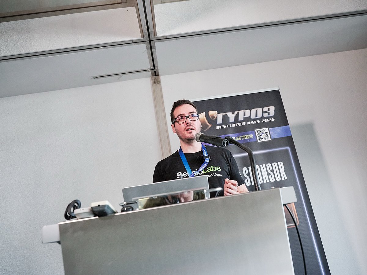 A person speaking at a podium during TYPO3 Developer Days 2026, with a microphone and laptop, and a sponsor banner in the background.