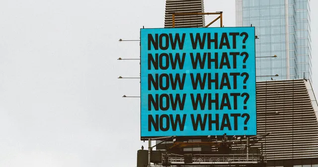 Blue sign on a building with several Now What? letters