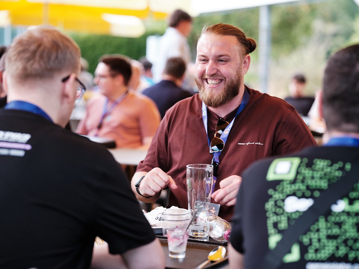 A bearded man in a brown shirt smiles and chats with two others at an outdoor gathering, with drinks and glasses on the table.