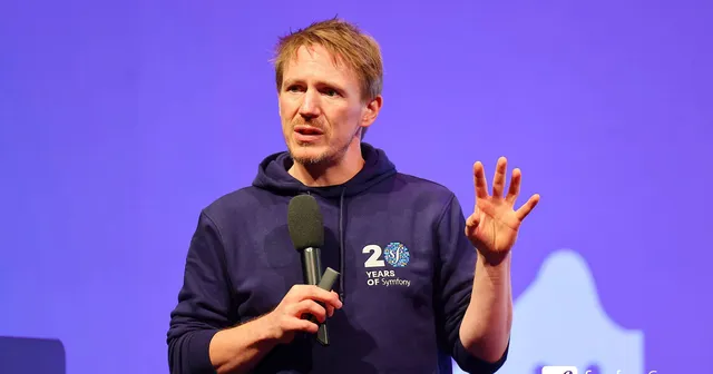 Nicolas Grekas with a mic in his right hand raising his left hand on stage at SymfonyCon Amsterdam 2025