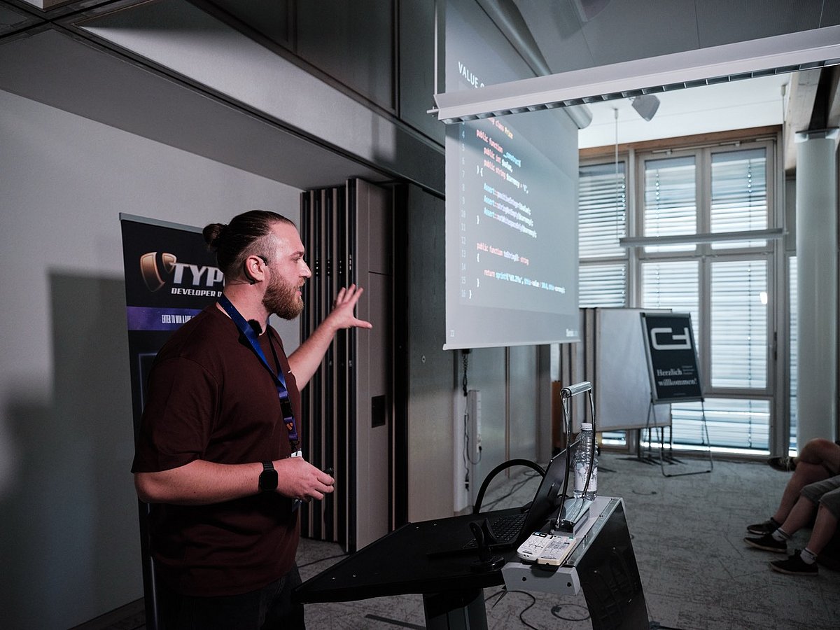 A man giving a presentation on coding, gestures towards a projected screen displaying code in a modern conference room.
