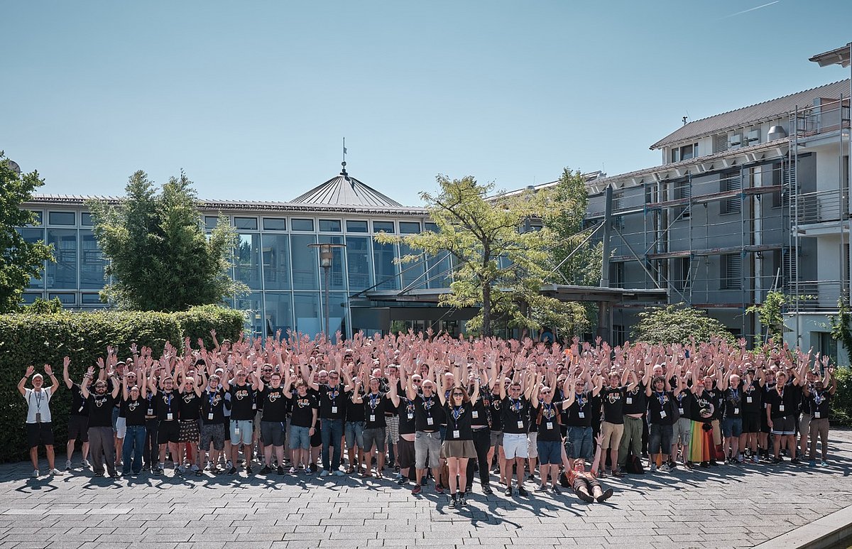 A large group of people wearing matching black shirts raise their arms in celebration outside a modern glass building under a clear blue sky.